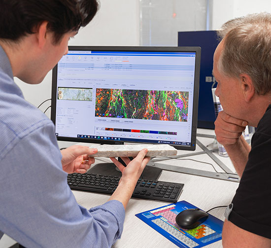 Two men looking at a computer screen comparing a core geological sample with a mineral map of it on screen.